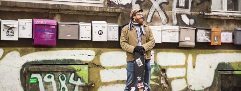 Jan Bratenstein steht mit Gitarre vor einem Haus mit vielen Briefkästen