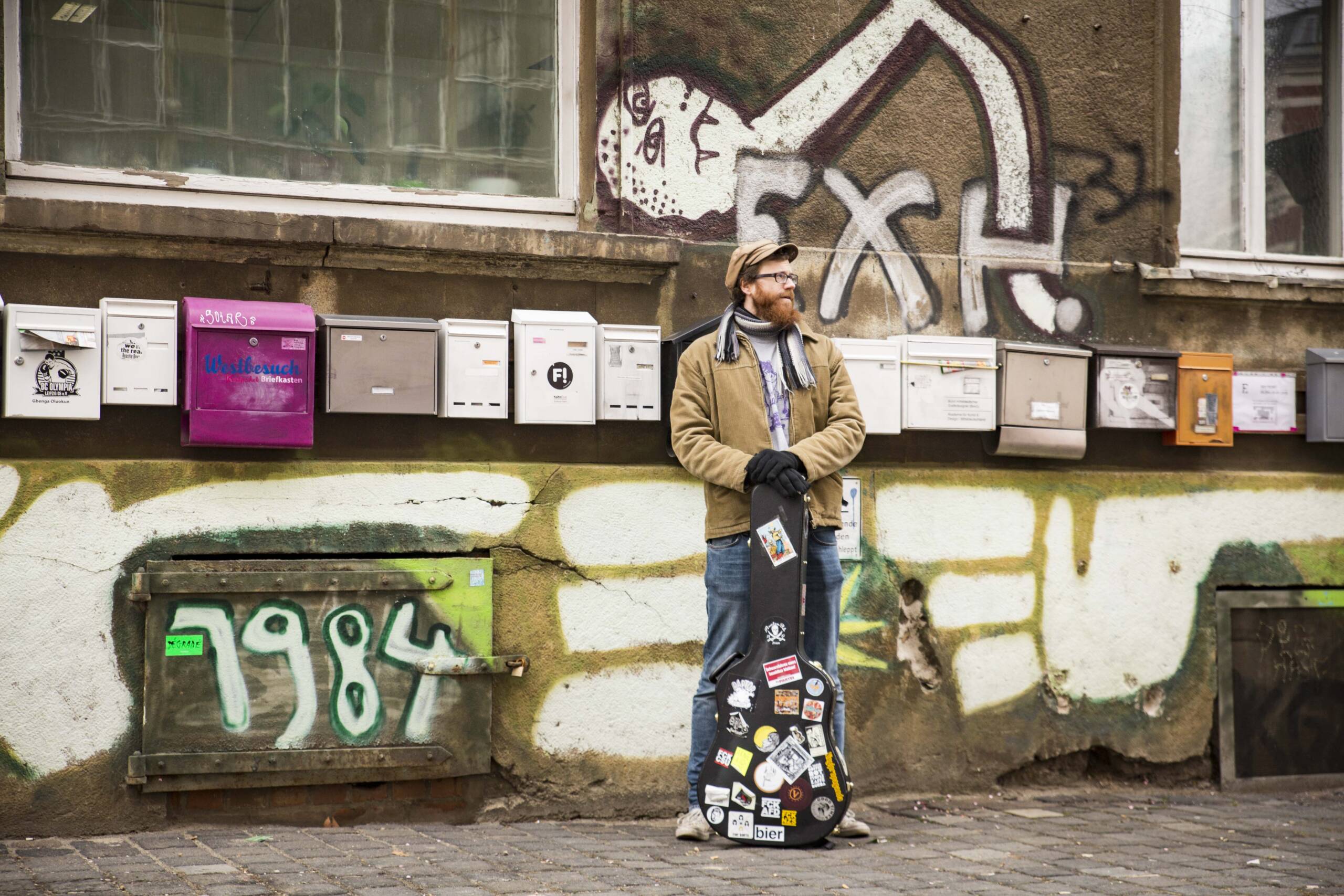 Jan Bratenstein steht mit Gitarre vor einem Haus mit vielen Briefkästen